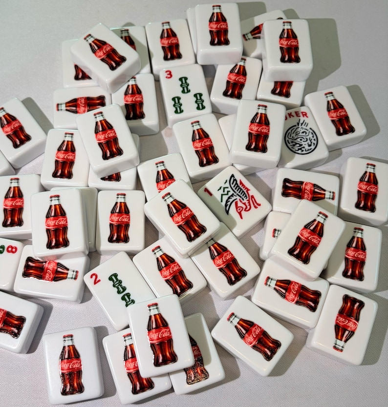 May include: A collection of white rectangular mahjong tiles featuring a Coca-Cola bottle graphic on the front. Some tiles display the red Coca-Cola logo, while others show green numerical symbols or a black and white graphic of a dragon. The tiles are scattered.