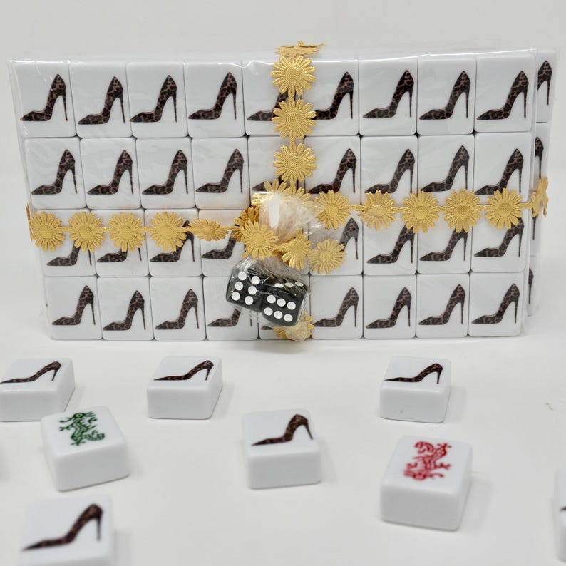 May include: A set of white Mahjong tiles with a high-heeled shoe design in a leopard print. The tiles are wrapped with a gold ribbon and flower accents. A black and white die is included. The tiles are for the game of Mahjong.