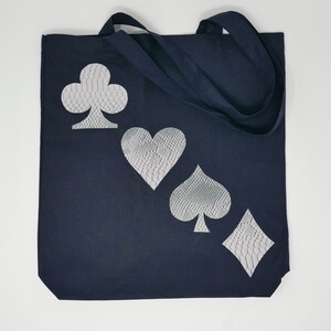 Puede incluir: Una bolsa de lona azul oscuro con un diseño blanco de los cuatro palos de las cartas de juego: tréboles, corazones, picas y diamantes. Los palos están estilizados con un patrón texturizado.