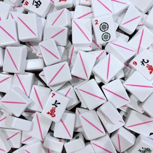 Puede incluir: Una colecci&oacute;n de fichas de mahjong rectangulares blancas esparcidas. Muchas fichas presentan una &uacute;nica l&iacute;nea diagonal rosa, mientras que otras muestran caracteres chinos rojos o negros y s&iacute;mbolos num&eacute;ricos, incluyendo los n&uacute;meros 2, 7 y 9.
