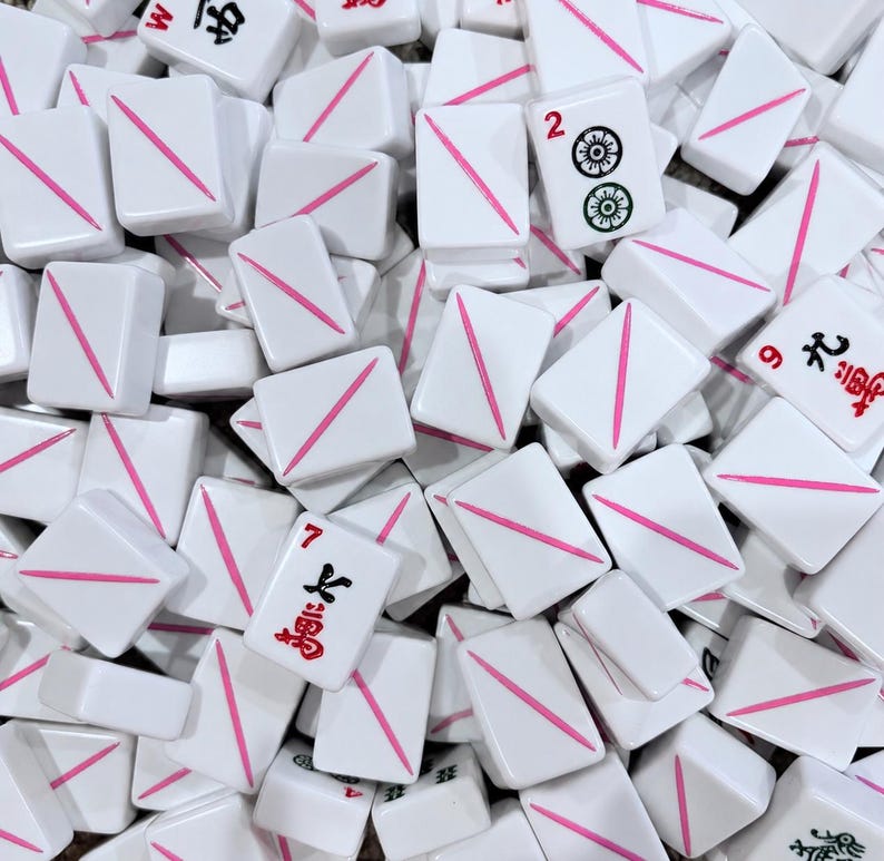 Puede incluir: Una colecci&oacute;n de fichas de mahjong rectangulares blancas esparcidas. Muchas fichas presentan una &uacute;nica l&iacute;nea diagonal rosa, mientras que otras muestran caracteres chinos rojos o negros y s&iacute;mbolos num&eacute;ricos, incluyendo los n&uacute;meros 2, 7 y 9.