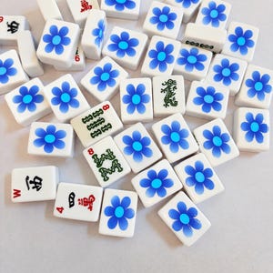 May include: A collection of white rectangular mahjong tiles scattered across a surface. Most tiles feature a blue flower design with a light blue center. Several tiles display traditional Chinese characters in red or black, and one tile features a green dragon illustration.