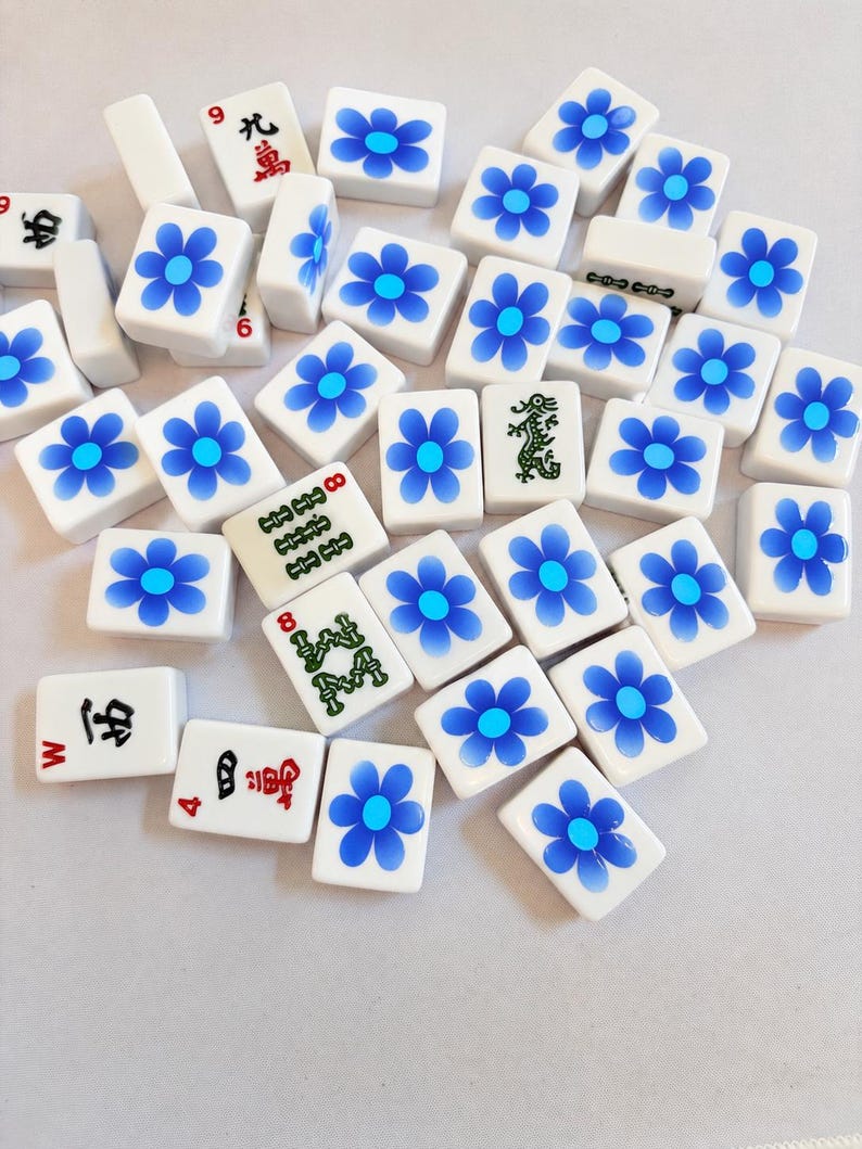 May include: A collection of white rectangular mahjong tiles scattered across a surface. Most tiles feature a blue flower design with a light blue center. Several tiles display traditional Chinese characters in red or black, and one tile features a green dragon illustration.