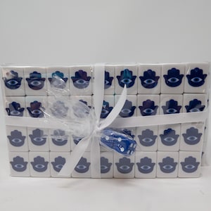 Könnte beinhalten: Eine Sammlung weißer quadratischer Fliesen, jede mit einer blauen Hamsa-Hand und einem bösen Blick-Design. Die Fliesen sind in durchsichtigem Zellophan verpackt und mit einem weißen Band gebunden. Ein blauer Würfel ist enthalten.