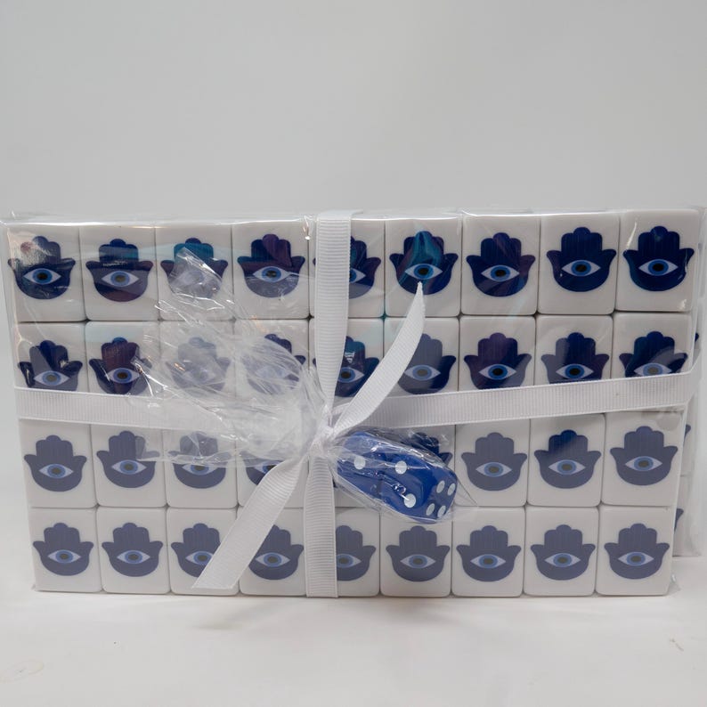 May include: A collection of white square tiles, each adorned with a blue Hamsa hand and an evil eye design. The tiles are wrapped in clear cellophane and tied with a white ribbon. A blue dice is included.