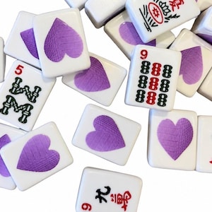May include: A collection of white rectangular mahjong tiles scattered across a surface. Several tiles feature a purple heart design, while others display traditional Chinese characters in black and red ink, including the numbers five, nine, and four.