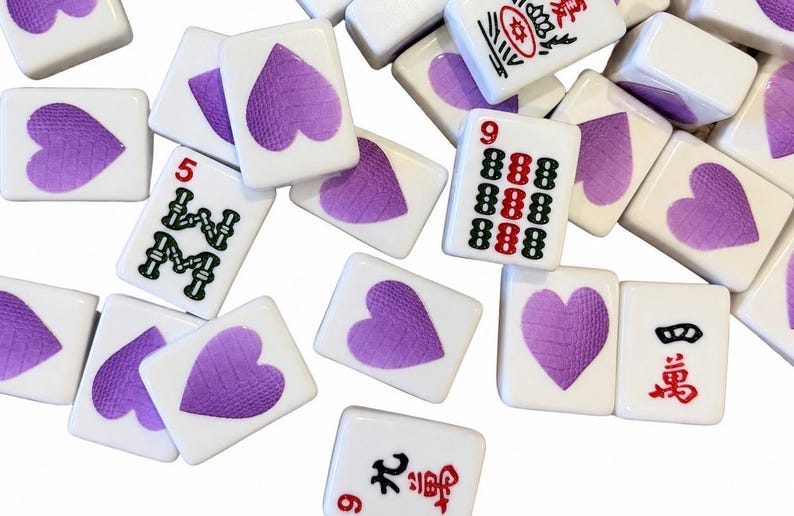 May include: A collection of white rectangular mahjong tiles scattered across a surface. Several tiles feature a purple heart design, while others display traditional Chinese characters in black and red ink, including the numbers five, nine, and four.