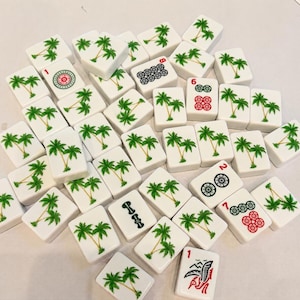 May include: A collection of white rectangular mahjong tiles scattered across a surface. Most tiles feature a green palm tree illustration, while others display red and black numerical symbols, floral patterns, and a red bird-like graphic.
