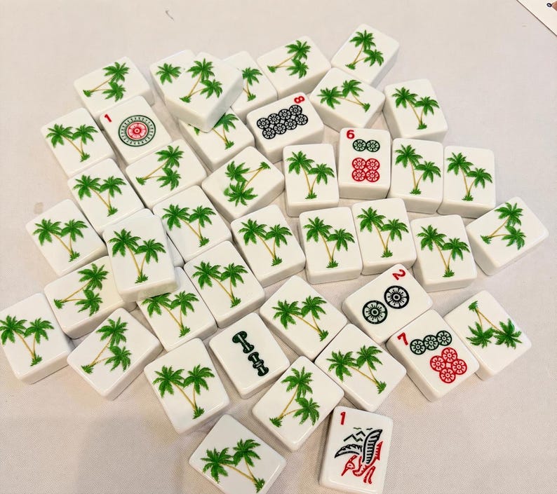 May include: A collection of white rectangular mahjong tiles scattered across a surface. Most tiles feature a green palm tree illustration, while others display red and black numerical symbols, floral patterns, and a red bird-like graphic.