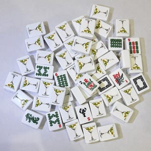 May include: A collection of white rectangular mahjong tiles featuring various illustrations, including martini glasses with olives, green Chinese characters, red numerical symbols, and black and red decorative patterns scattered across a plain surface.