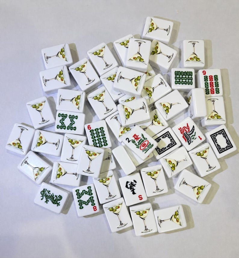 May include: A collection of white rectangular mahjong tiles featuring various illustrations, including martini glasses with olives, green Chinese characters, red numerical symbols, and black and red decorative patterns scattered across a plain surface.