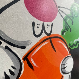 May include: A white bunny with a pink nose and black eyes is holding a carrot in its paws. The bunny is painted on a black background with green and orange accents.