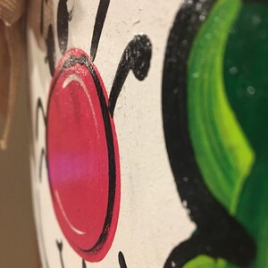 May include: Close-up of a white painted wooden ornament with a red nose, black eyes, and green leaves. The ornament is painted with a whimsical style.