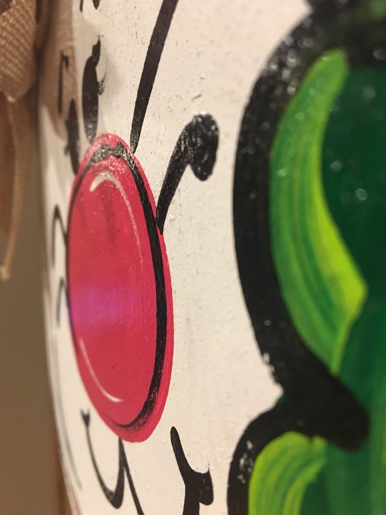 May include: Close-up of a white painted wooden ornament with a red nose, black eyes, and green leaves. The ornament is painted with a whimsical style.