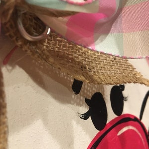 May include: Close-up of a pink and white plaid ribbon tied around a burlap bow. The ribbon is tied around a white surface with a black and red painted design.