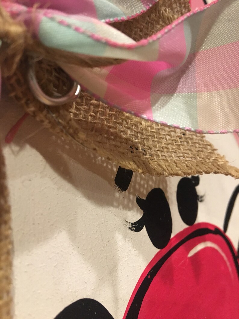May include: Close-up of a pink and white plaid ribbon tied around a burlap bow. The ribbon is tied around a white surface with a black and red painted design.