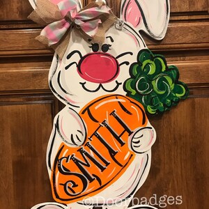 May include: White bunny door hanger with pink ears and a pink nose. The bunny is holding an orange carrot with the name "Smith" written on it. The bunny is wearing a pink and white plaid bow.