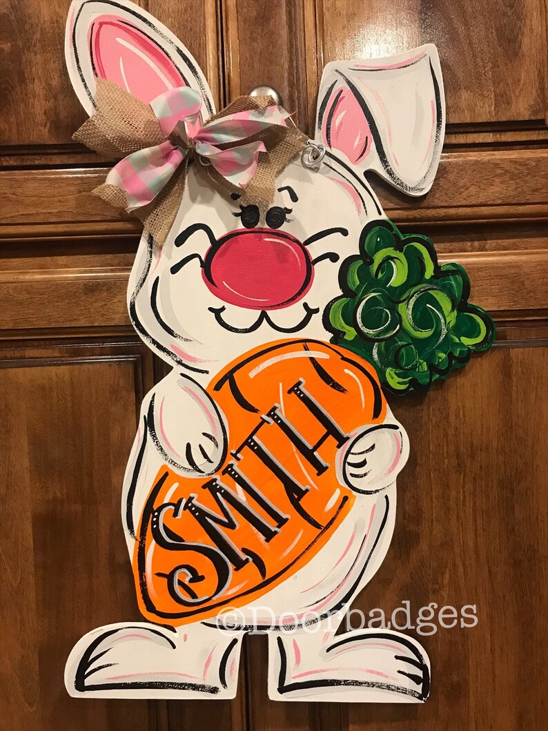 May include: White bunny door hanger with pink ears and a pink nose. The bunny is holding an orange carrot with the name "Smith" written on it. The bunny is wearing a pink and white plaid bow.