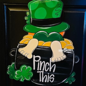 May include: A wooden door hanger featuring a leprechaun with a green hat and orange beard, sitting in a black pot filled with gold coins. The pot has the text "Pinch This" written on it.