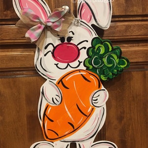 May include: White bunny door hanger with pink ears, a pink and white bow, and a large orange carrot. The bunny is holding a bunch of green leaves.