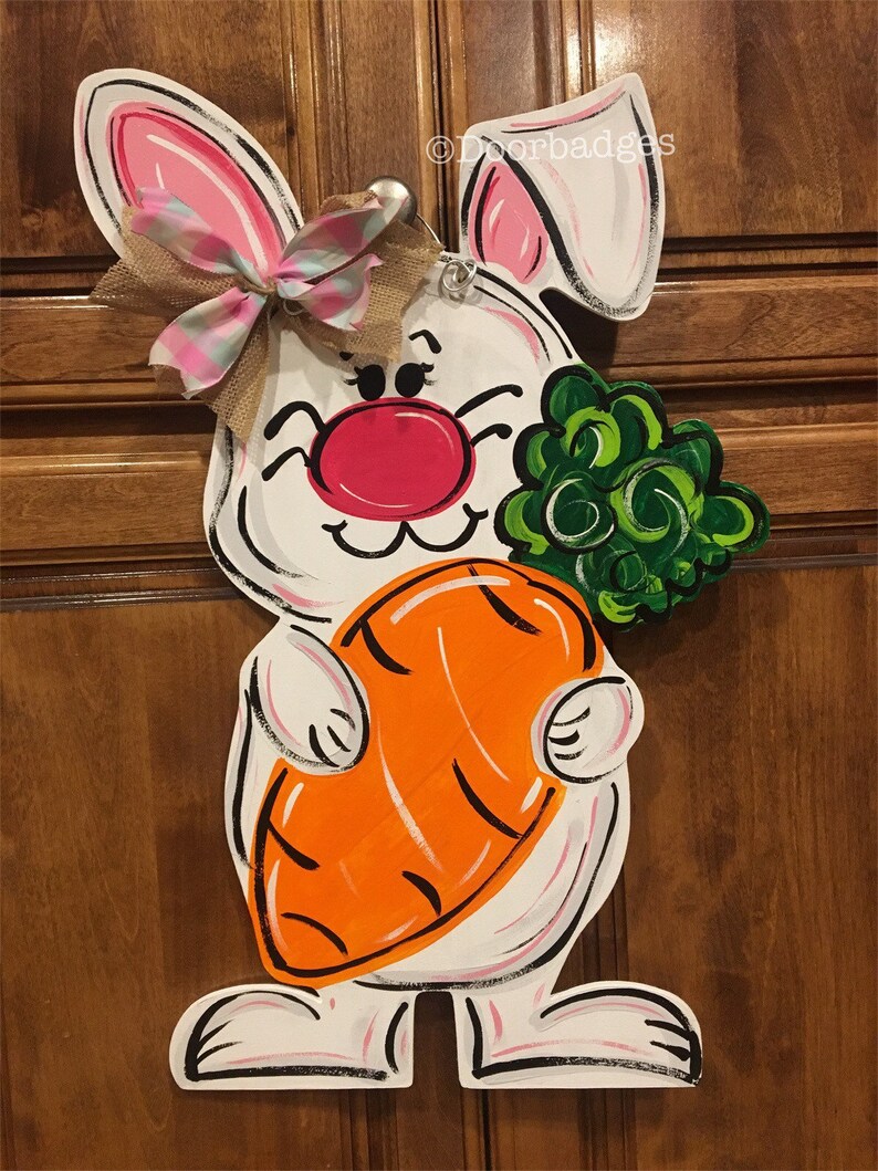 May include: White bunny door hanger with pink ears, a pink and white bow, and a large orange carrot. The bunny is holding a bunch of green leaves.