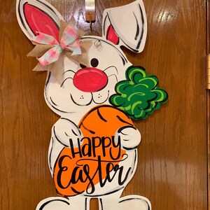 May include: White bunny rabbit holding a carrot with the words "Happy Easter" written on it. The bunny has a pink nose, green clover, and a pink and white bow.