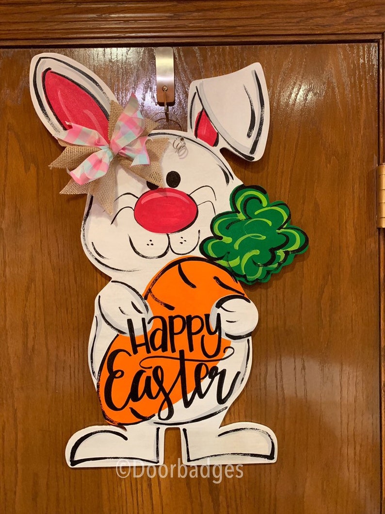 May include: White bunny rabbit holding a carrot with the words "Happy Easter" written on it. The bunny has a pink nose, green clover, and a pink and white bow.