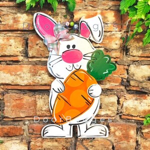May include: White bunny rabbit holding a carrot with green leaves. The bunny has pink ears, a pink nose, and a pink bow. The carrot is orange with brown spots.