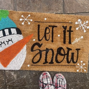May include: A brown coir doormat with the text "Let it snow" in black script. A white snowman with a blue hat and orange carrot nose is on the left side of the mat. There are white snowflake designs on the mat.