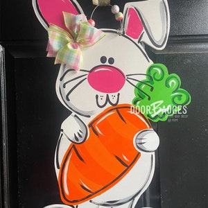 May include: A white bunny holding a carrot with green leaves. The bunny has pink ears, a pink nose, and a pink bow with green and white checkered ribbon. The bunny is standing on a black background.