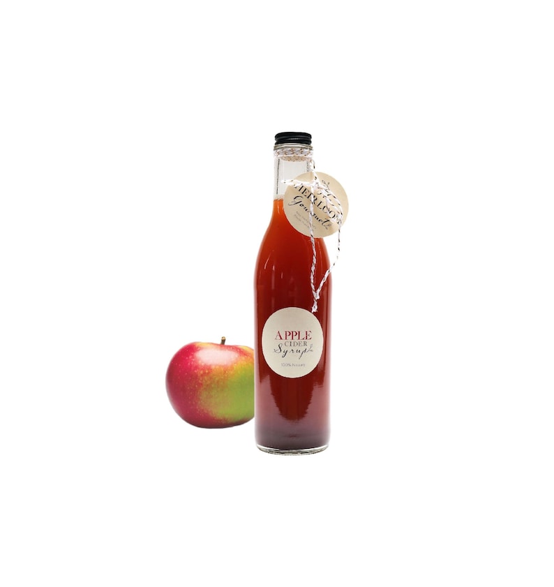 May include: A glass bottle of apple cider syrup with a black cap and a white label that says "APPLE CIDER SYRUP 100% Natural". A red apple is in the foreground.