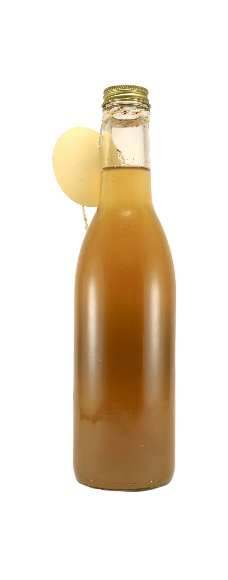 Puede incluir: Una botella de vidrio con una tapa dorada y un l&iacute;quido marr&oacute;n claro en su interior. La botella tiene una cuerda atada alrededor del cuello con una peque&ntilde;a etiqueta redonda amarilla.