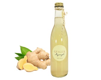 May include: A clear glass bottle filled with light yellow ginger syrup, featuring a round label that reads GINGER Syrup 100% Nature. The bottle has a gold cap and is tied with a thin string. Beside it sits a fresh ginger root with green leaves.