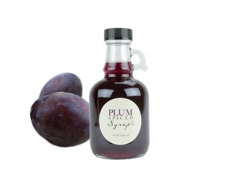 May include: A glass bottle with a black lid and a handle filled with a dark red liquid. The bottle has a white label that says "Plum Spiced Syrup" and "100% Natural". Three purple plums are in the foreground.