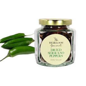 May include: A clear glass jar with a gold lid filled with dried serrano peppers. A white label on the jar reads "The Heirloom Gourmet" and "Dried Serrano Peppers 100% Natural." Four fresh green serrano peppers are to the left of the jar.
