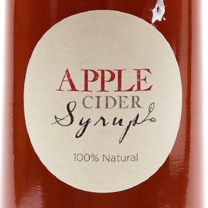 May include: A round, white label with the text "APPLE CIDER Syrup" in red and gray lettering. The label also says "100% Natural" in gray lettering. The label is on a brown glass bottle.
