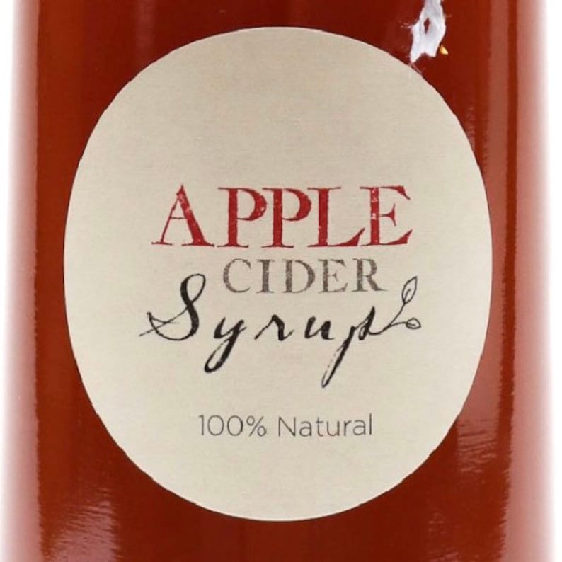 May include: A round, white label with the text "APPLE CIDER Syrup" in red and gray lettering. The label also says "100% Natural" in gray lettering. The label is on a brown glass bottle.