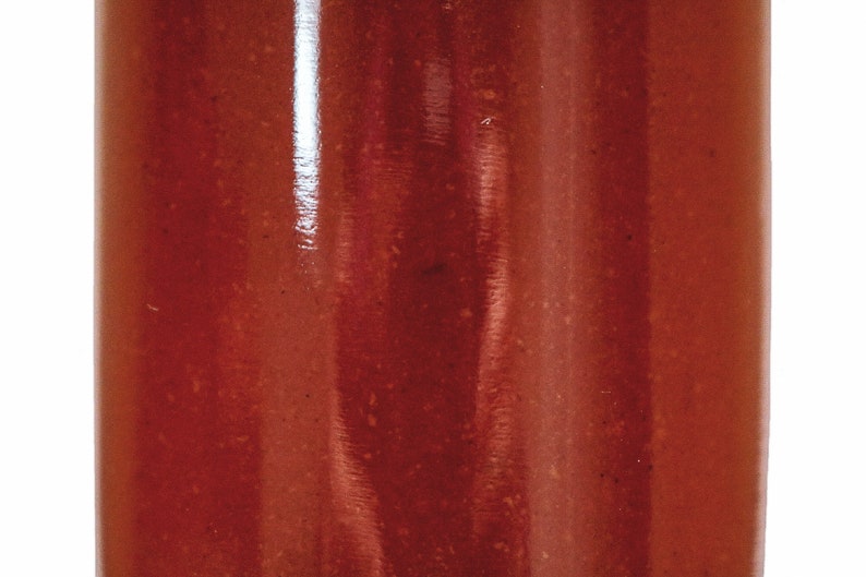 May include: A close-up of a glass jar filled with a thick, red liquid. The liquid has a slightly textured surface and appears to be a sauce or condiment.
