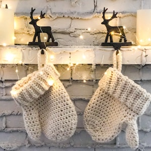 May include: Two cream-coloured crocheted stockings hanging from a white brick fireplace mantel with black reindeer stocking holders and fairy lights.