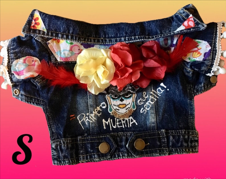 May include: A blue denim jacket with a white embroidered skull and the text "Primero Muerta que Sencilla!"  The jacket has a white lace trim and a red feather.  The jacket is decorated with colorful flowers.