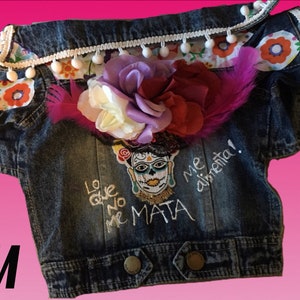 May include: A blue denim jacket with white pom pom trim and a floral design. The jacket has an embroidered design with the text "Lo que no me mata me alimenta!" and a skull with flowers.