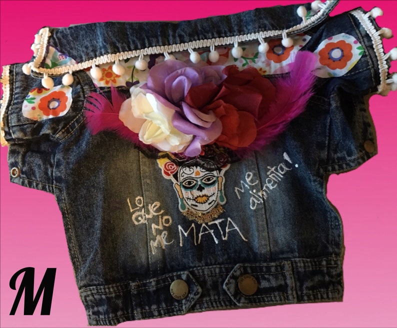 May include: A blue denim jacket with white pom pom trim and a floral design. The jacket has an embroidered design with the text "Lo que no me mata me alimenta!" and a skull with flowers.