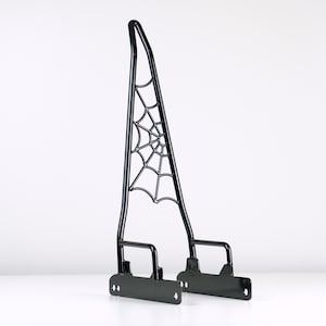 May include: Black metal motorcycle sissy bar with a spiderweb design. The sissy bar has two mounting brackets.