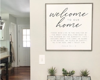 May include: A framed sign with the text "Welcome to our home" and a bible verse from Psalm 91:1-2. The sign is hanging on a light gray wall above a wooden table with a metal tray filled with succulents.