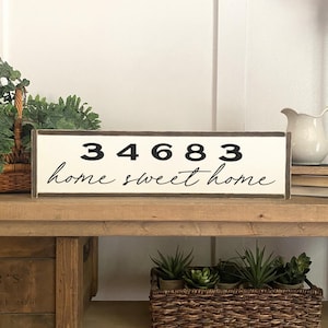 May include: A white wooden sign with a brown frame. The sign has black text that reads "34683 home sweet home".