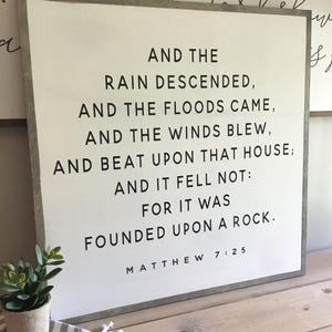 May include: A white wooden sign with black text that reads "And the rain descended, and the floods came, and the winds blew, and beat upon that house; and it fell not: for it was founded upon a rock. Matthew 7:25".