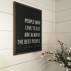 May include: A black framed sign with white text that reads "People who love to eat are always the best people." The text "Julia Child" is written in cursive below the quote. The sign is hanging on a white wall with white shiplap boards.