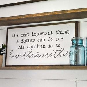 May include: White wooden sign with black text that reads "the most important thing a father can do for his children is to love their mother". The sign is sitting on a shelf with two clear glass jars with blue lids.