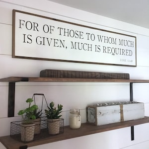 May include: A wooden sign with the text "FOR OF THOSE TO WHOM MUCH IS GIVEN, MUCH IS REQUIRED" and the scripture reference "LUKE 12:48". The sign is hanging on a white wall above a wooden shelf.