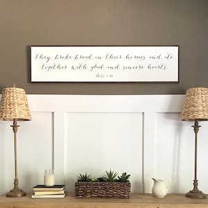 May include: A wooden sign with a white background and black text. The text reads "They broke bread in their homes and ate together with glad and sincere hearts. Acts 2:46".